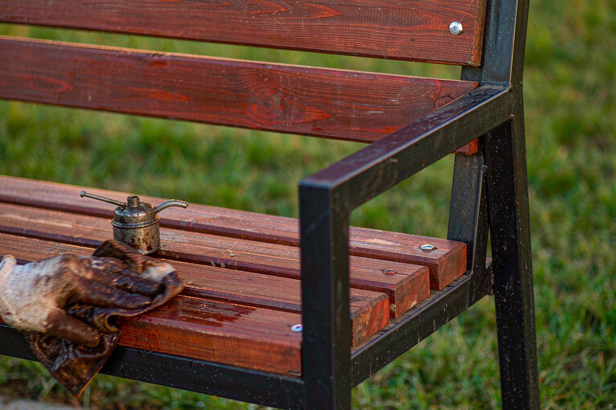 Gloved hand wiping oil onto reddish-brown hardwood park bench slats with a cloth and oil can