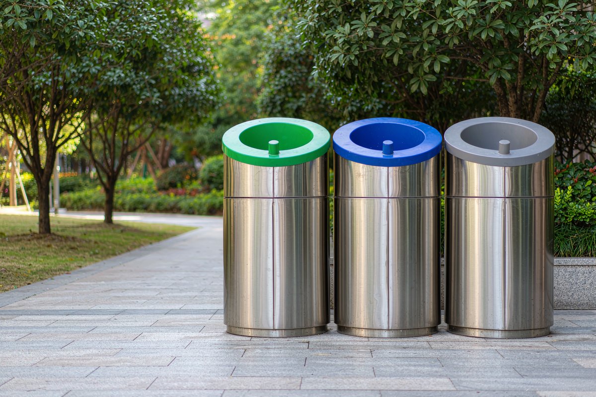 Three stainless steel waste-sorting bins with colour-coded lids standing side by side on a park plaza